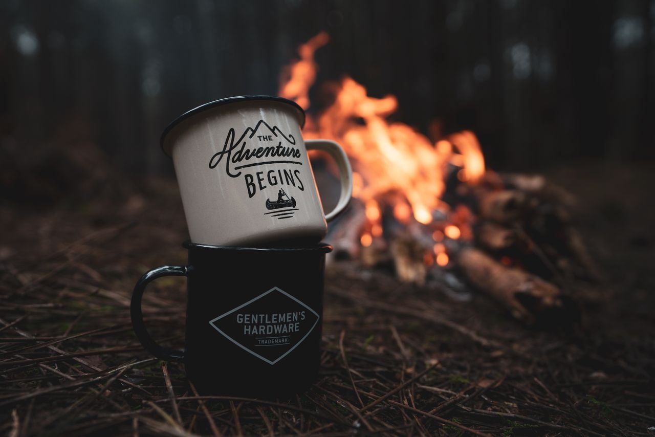 Two mugs with inspirational quotes stacked near a campfire in a tranquil forest environment.