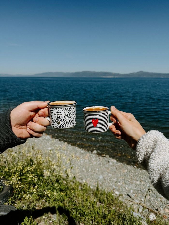 Two hands holding decorative mugs in a celebratory toast by a serene lakeside with a mountain backdrop.