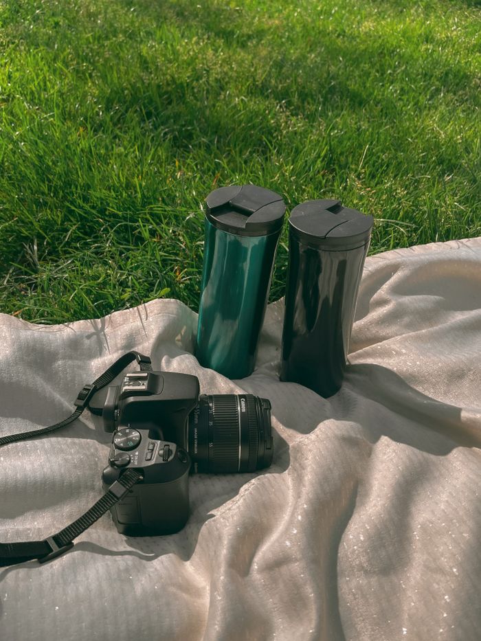 A DSLR camera and two coffee mugs on a blanket outdoors in green grass.